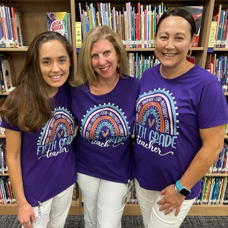 The 2022-2023 Skaggs 5th grade team standing and smiling in the Skaggs library by a bookshelf. They are wearing their purple 5th grade team shirts and white jeans.