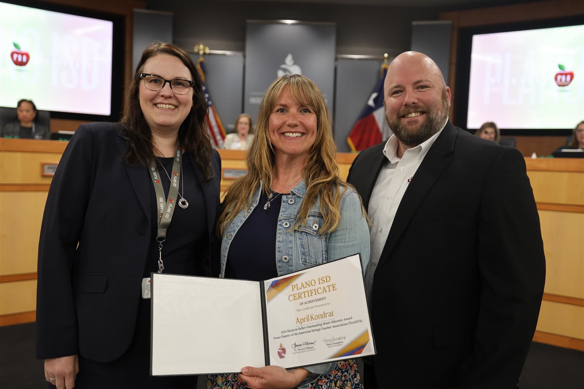 Three individuals in front of the Plano ISD Board Dais, one holding a certificate of accomplishment