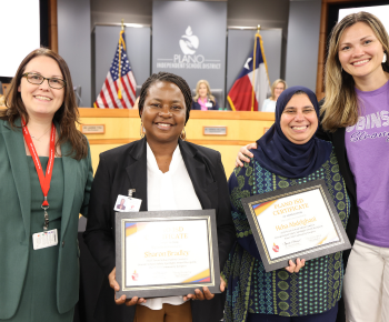   Four individuals posing in front of the Plano ISD Board of Trustees dais