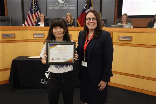Two individuals posing in front of the Plano ISD Board of Trustees Dais 