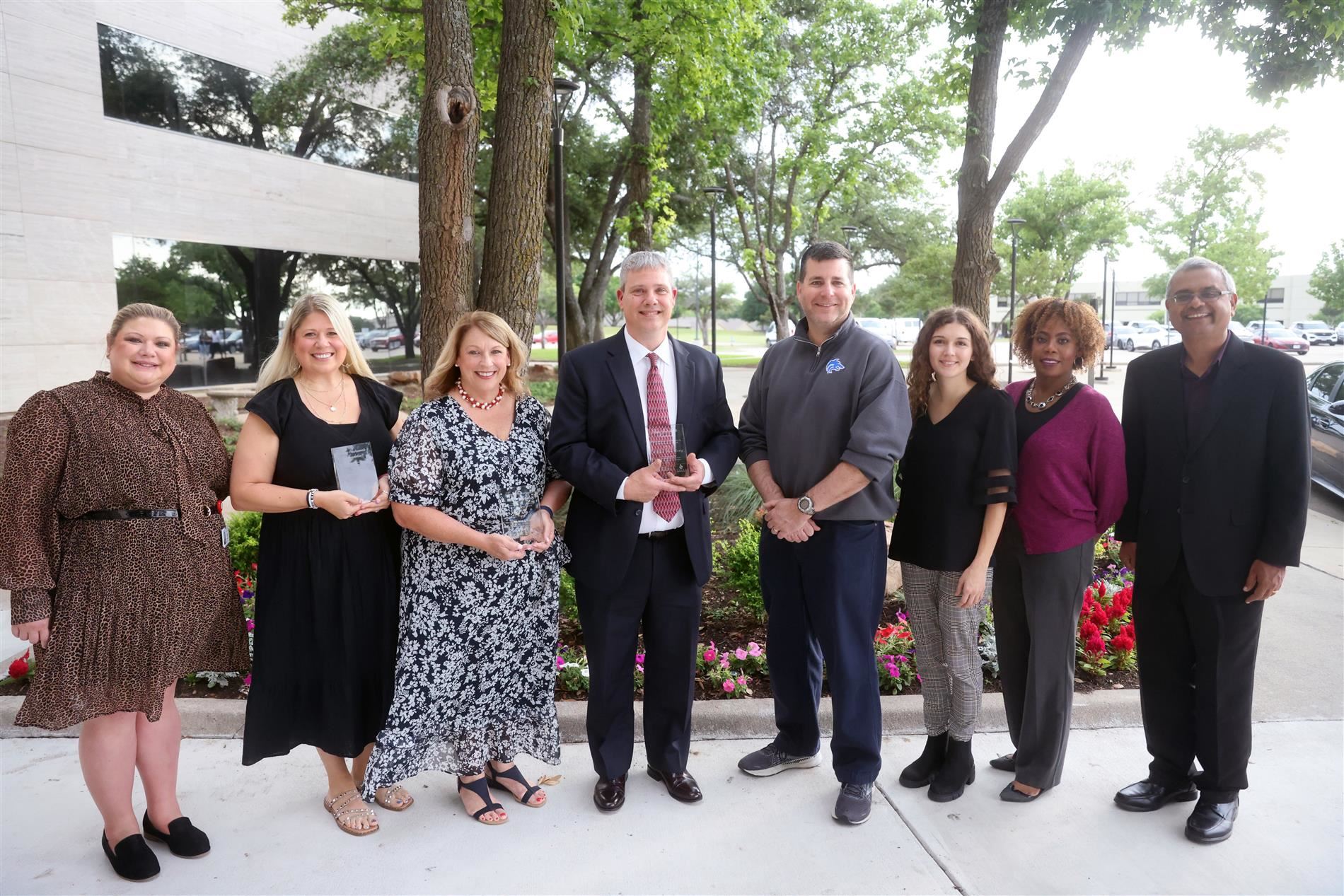 a group photo of 8 individuals in an outside setting. 3 of which are holding awards.