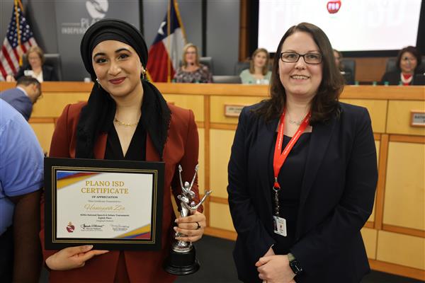 Two individuals posing in front of the Plano ISD Board Dais