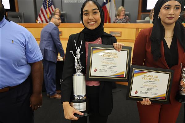 Two individuals posing in front of the Plano ISD Board Dais
