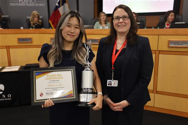 Two individuals posing in front of the Plano ISD Board Dais