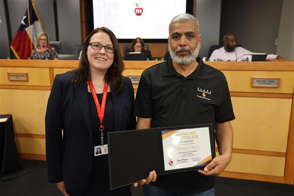 Two individuals posing in front of the Plano ISD Board Dais