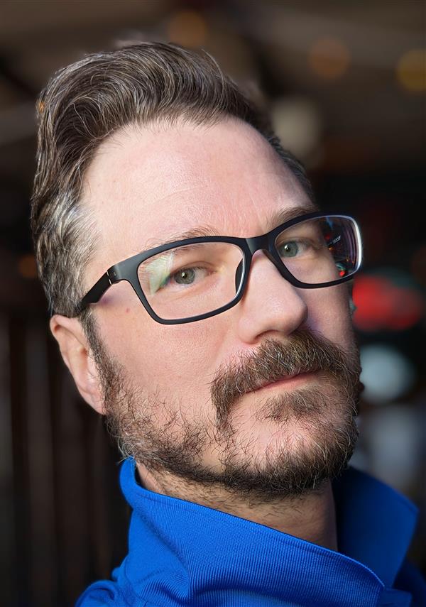 This is a headshot of a white man with brown hair, and a brown goatee. He is wearing black-framed glasses and a blue polo shirt. He is Mrs. Hoffmann's husband.